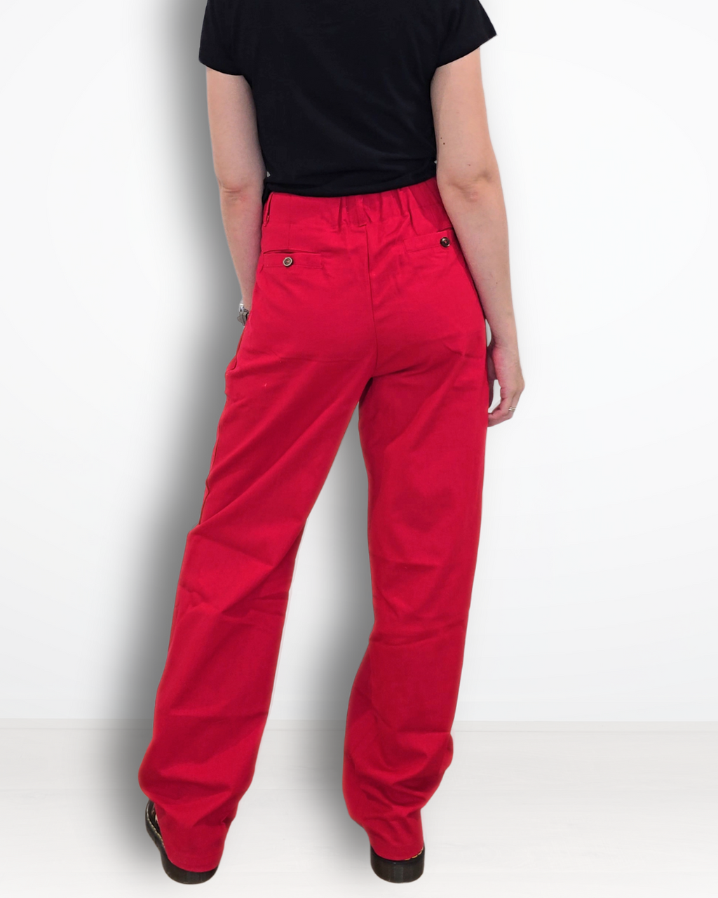A woman viewed from behind wearing bright red straight-leg pants, a black short-sleeve t-shirt, and black shoes, standing against a plain white studio background.