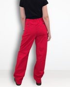A woman viewed from behind wearing bright red straight-leg pants, a black short-sleeve t-shirt, and black shoes, standing against a plain white studio background.