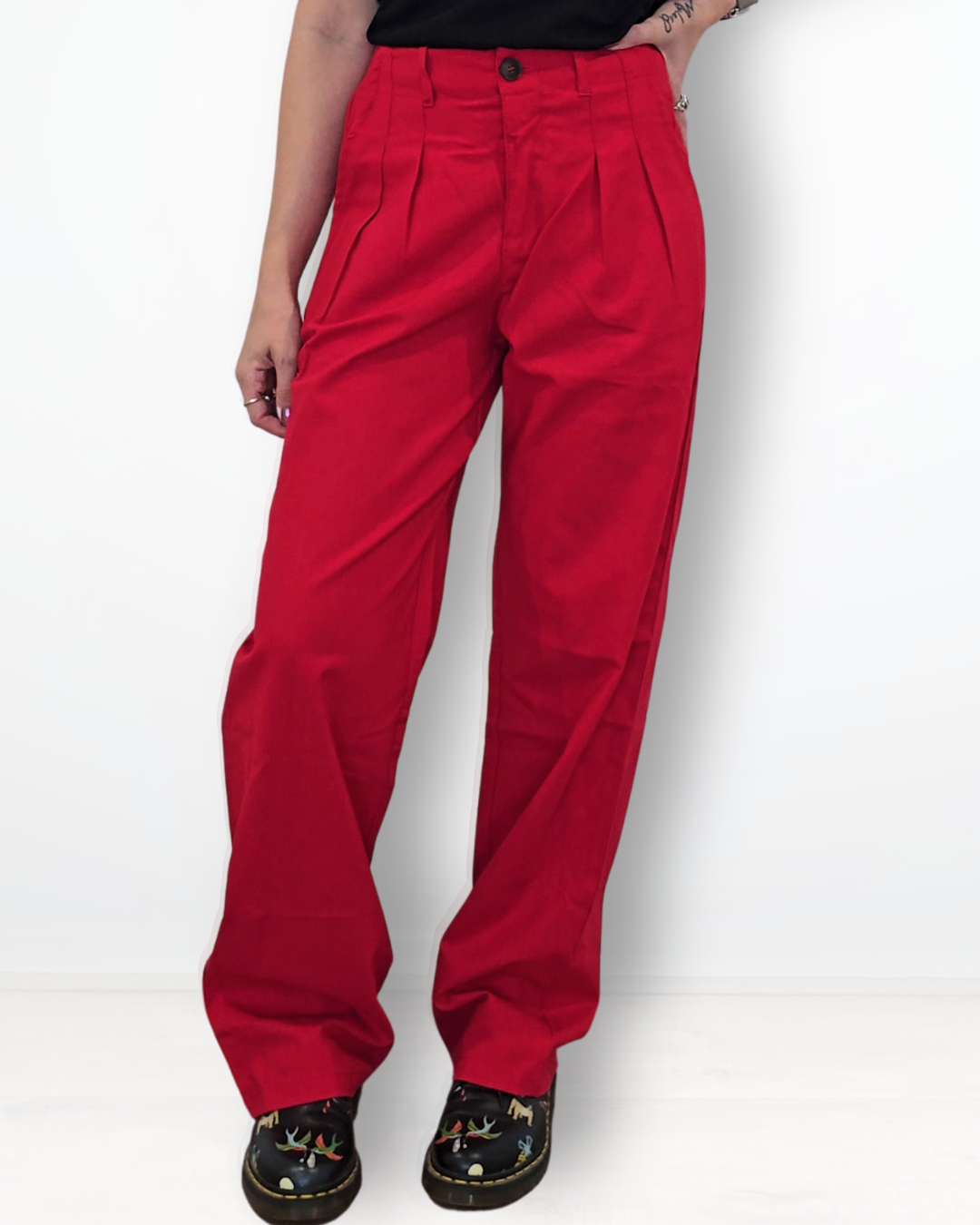 A woman modeling a pair of bright red high-waisted, wide-leg pleated trousers, paired with a black t-shirt and black shoes, against a plain white background.
