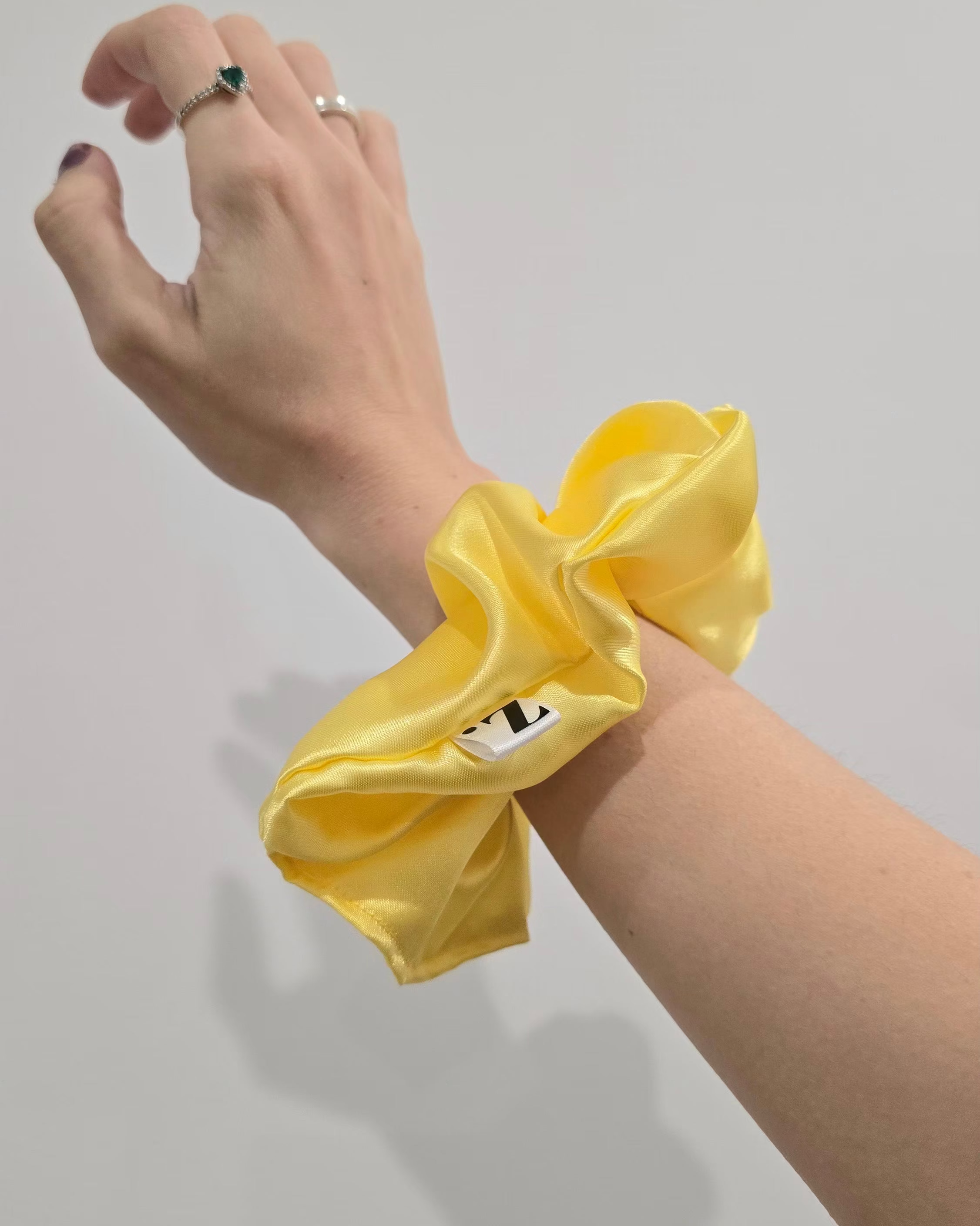 A bright yellow satin hair scrunchie worn on a person's wrist, shown against a plain light gray background.