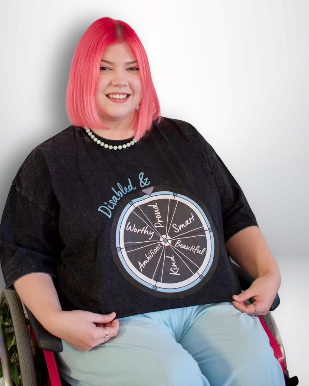 Person with pink hair wearing a black t-shirt with text and graphics, sitting in a wheelchair against a white background.