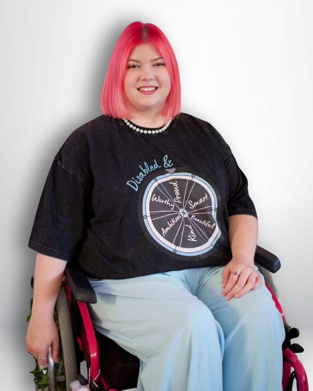 Person with pink hair wearing a black t-shirt with a graphic design, sitting in a wheelchair against a plain background.