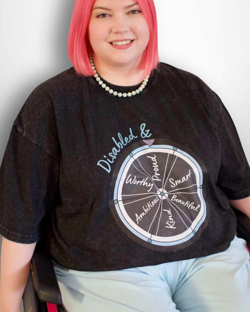 Person with pink hair wearing a black t-shirt with text and graphics, sitting in a wheelchair on a white background.
