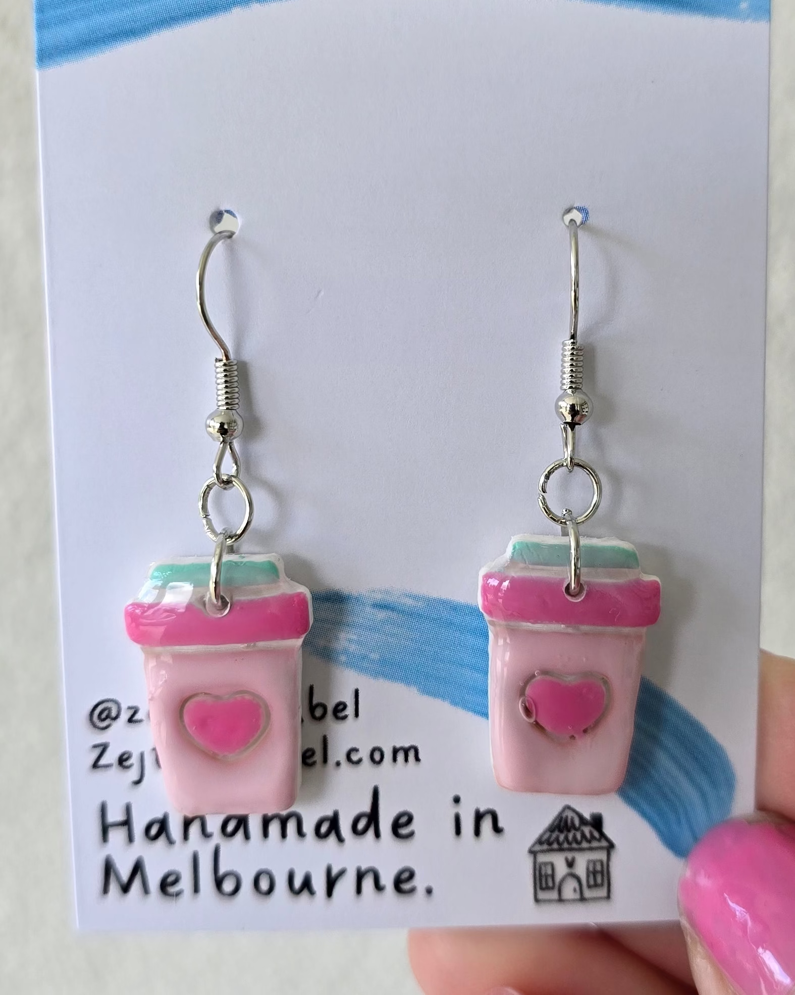 Pink coffee cup earrings on a 'Zejt' branded card held by a hand.