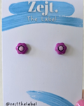 Purple earrings on a 'Zejt. The Label' card held by a hand with pink nail polish.
