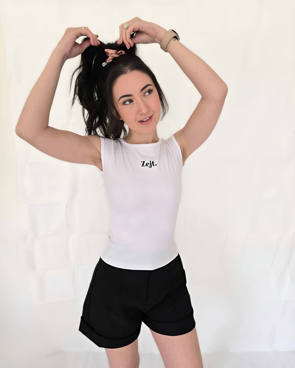 Woman wearing a white tank top with 'Zeit' logo and black shorts on a plain background, cute pink hair accessory scrunchie